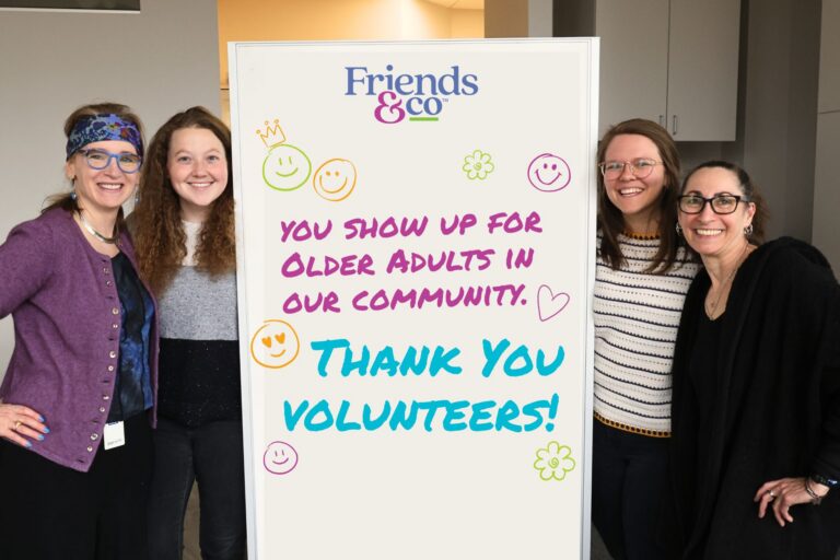 Friends & Co programming staff posing beside a marker board with the words written out "You show up for older adults in our community. Thank you Volunteers!"